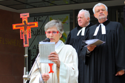 A religious leader in white vestments reads a prayer with several other religious leaders standing behind her in black robes.