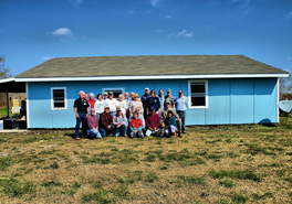 A group of people sitting and standing together for a photo outside of a now blue-colored house they constructed.