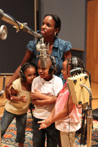 A woman with a group of three children, recording a song into a microphone.